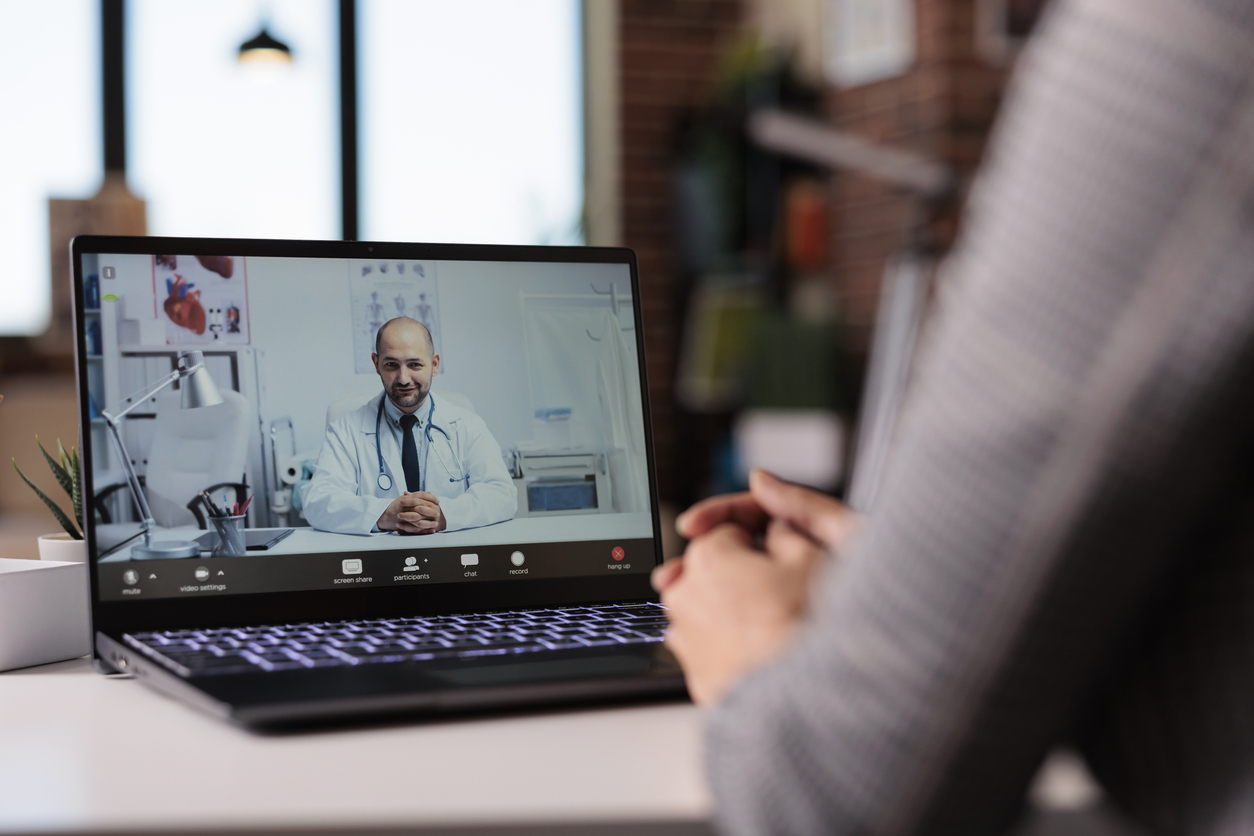 Eine Person sitzt vor einem Laptop und am Bildschirm ist ein Arzt zu sehen - eine telemedizinische Konsultationsszene.