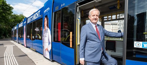 Ein steht in der Tür einer geöffneten Straßenbahn - mit einem Bein noch in der Straßenbahn, mit dem anderen bereits am Trottoir.