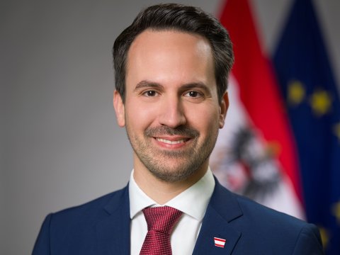 Ein Porträt von Bildungsminister Christoph Wiederkehr mit freundlichem Blick - im Hintergrund sind verschwommen die Flaggen von Österreich und der Europäischen Union zu sehen.