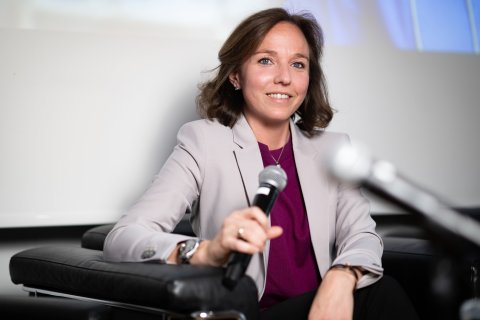 Ein Frau sitzt in einem Fauteuil, hält ein Mikrofon in der Hand und lächelt in die Kamera.
