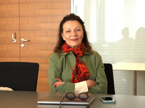 Eine Frau mit rotem Schal sitzt an einem Tisch mit nach vorne verschränkten Armen und blickt direkt in die Kamera.