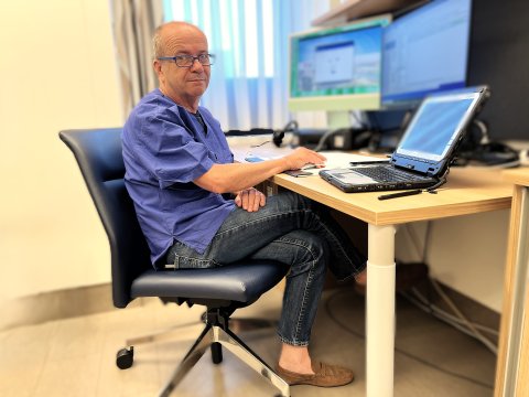 Ein Mann sitzt in einem Sessel vor einem Schreibtisch, auf dem ein Laptop und zwei zusätzliche Bildschirme stehen.
