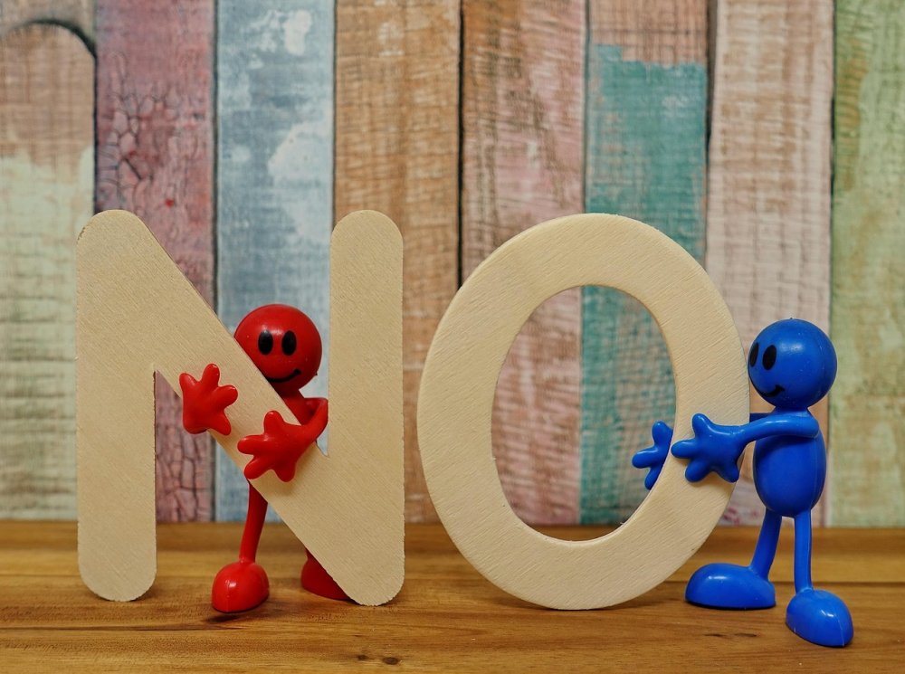 Auf dem Foto ist in hölzernen Buchstaben das Wort "No", also Nein, zu sehen, die von jeweils einer Spielzeugfigur, eine in blau, die andere in rot, gehalten werden.