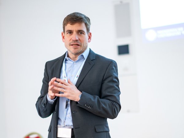 Ein Mann steht bei einem Vortrag und spricht, während er beide Hände vorne zusammenführt beim Sprechen. 