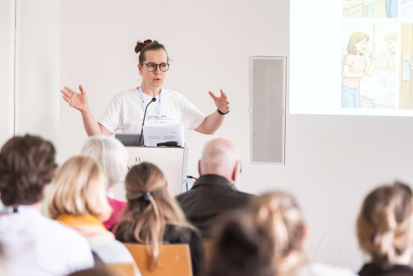Eine Frau hält gerade einen Vortrag, spricht und hält beide Hände und Arme vor sich, so als würde sie ein Längenmaß beschreiben.