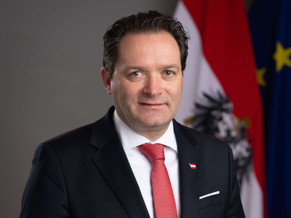 Der Bundesminister für Landwirtschaft Norbert Totschnig blickt in die Kamera - hinter ihm sieht man Teile der Fahnen von Österreich und der EU.